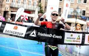 Athletin läuft jubelnd durch das Zielband des DATAGROUP Triathlon Nürnberg auf dem Hauptmarkt, im Hintergrund Zuschauer und Banner mit DATAGROUP-Logo.