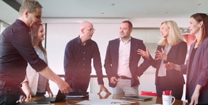 Gruppe von sechs Personen in einem modernen Konferenzraum in angeregter Unterhaltung. Zwei Männer und vier Frauen stehen um einen Tisch mit Laptops und Dokumenten, lächeln und diskutieren miteinander