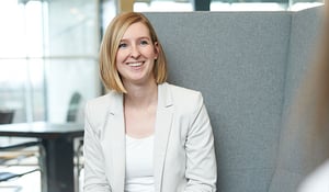 Nadine Schwaiger in einem weißen Blazer sitzt vor einem grauen Hintergrund.