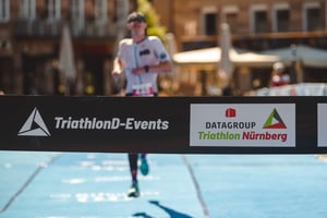 Zielband mit Logos von DATAGROUP Triathlon Nürnberg und TriathlonD-Events – im Hintergrund läuft ein Athlet ins Ziel.