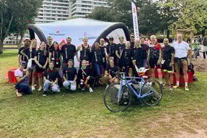 Gruppenfoto des DATAGROUP-Teams vor einem Pavillon beim Triathlon Nürnberg 2025 – Mitarbeitende in schwarzer Sportbekleidung mit DATAGROUP-Logo posieren gemeinsam, im Vordergrund ein Rennrad.