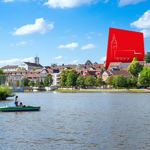 Blick über den Böblinger See mit Tretboot im Vordergrund, historischer Altstadt mit Wohnhäusern am Ufer und einer stilisierten roten Kirchturm-Silhouette unter blauem Himmel mit weißen Wolken